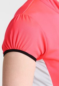 Koszulka sportowa z krótkim rękawem, wykonana z jasnoróżowej siateczkowej tkaniny, z marszczonym wzorem rękawów oraz czarnym kontrastowym obszyciem na rękawach.