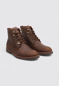 Botas de cuero marrón con puntas redondeadas, cordones en la parte delantera y ojales de latón. Suelas de goma texturizada con detalle de costura blanca.