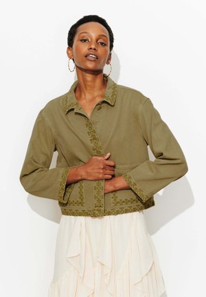 Femme aux cheveux courts portant de grandes boucles d'oreilles en anneau, une veste brodée vert olive et une jupe légère à volants sur fond blanc.