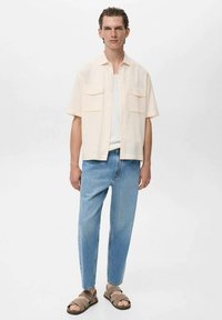 Kortærmet, lys ferskenfarvet skjorte med knapper og to brystlommer,parret med lyseblå tapered jeans og brune slidesandaler.