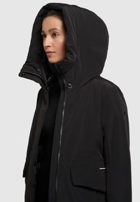 Sort vandtæt jakke med en stor hætte, lynlåslukning foran, trykknapper og sidelommer, der har en glat tekstur og en minimalisk design.