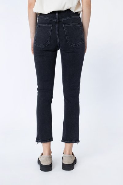 Baksidebilde av en person iført svarte avkortede jeans med frynsete kanter, hvit skjorte og beige sko mot en ensfarget hvit bakgrunn.