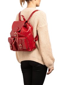 Zaino in pelle rossa con chiusura a cordoncino, tasche frontali e spallacci regolabili, indossato sopra un maglione in maglia beige. Texture liscia.