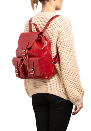 Sac à dos en cuir rouge avec une fermeture à cordon, des poches avant et des bretelles réglables, porté sur un pull en tricot beige. Texture lisse.