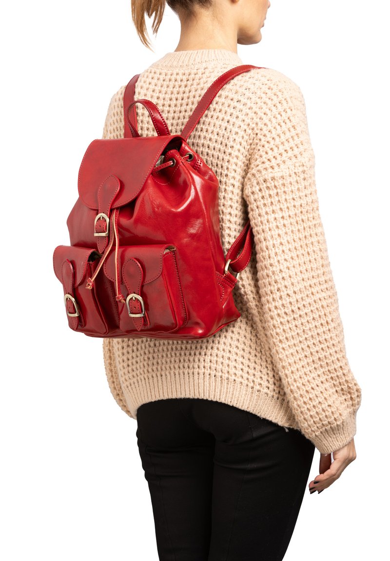 Zaino in pelle rossa con chiusura a cordoncino, tasche frontali e spallacci regolabili, indossato sopra un maglione in maglia beige. Texture liscia.