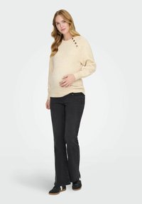 Maglione beige a maglia con scollatura rotonda, tre bottoni decorativi sulla spalla, abbinato a jeans neri a campana e sneakers nere.