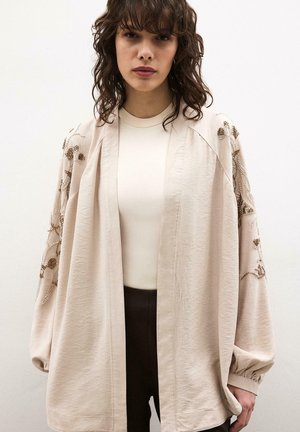 Femme aux cheveux bruns ondulés portant un cardigan ouvert crème brodé sur un haut blanc et un pantalon foncé, debout devant un fond uni.