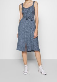 Vestido azul con lunares, escote cuadrado, botones frontales, cinturilla atada y dobladillo con volantes. Combinado con zapatillas blancas altas.