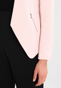 Ljusrosa kavaj gjord av mjukt tyg, med en sned fåll och en dragkedjestängning på vänster sida. Kombinerad med svarta byxor.