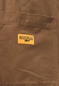 Bruine stof met een gestikt zakje met een klein rechthoekig geel label met de tekst "Service Works" en gereedschapsiconen onder de tekst.