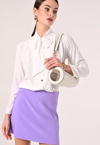 Weiße Bluse mit einem Schleifen-Detail, kombiniert mit einem fliederfarbenen Minirock. Eine runde Handtasche mit einem klaren, runden Fenster und goldenen Akzenten wird gehalten.