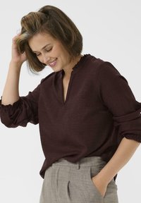 Blusa bordeaux con scollo a V, colletto arricciato e maniche arricciate. Indossata con pantaloni grigi chiari su misura. Tessuto testurizzato.