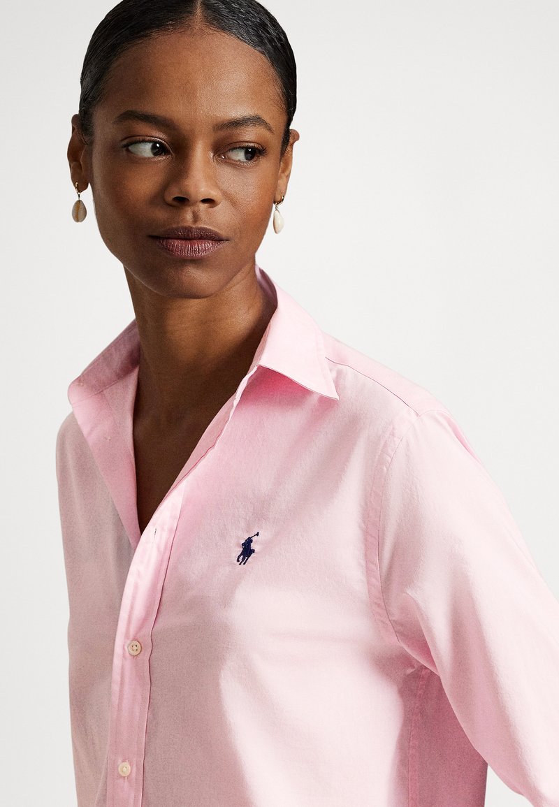 Donna con pelle scura indossa una camicia rosa chiaro con colletto e orecchini a conchiglia, guarda di lato su uno sfondo bianco semplice.