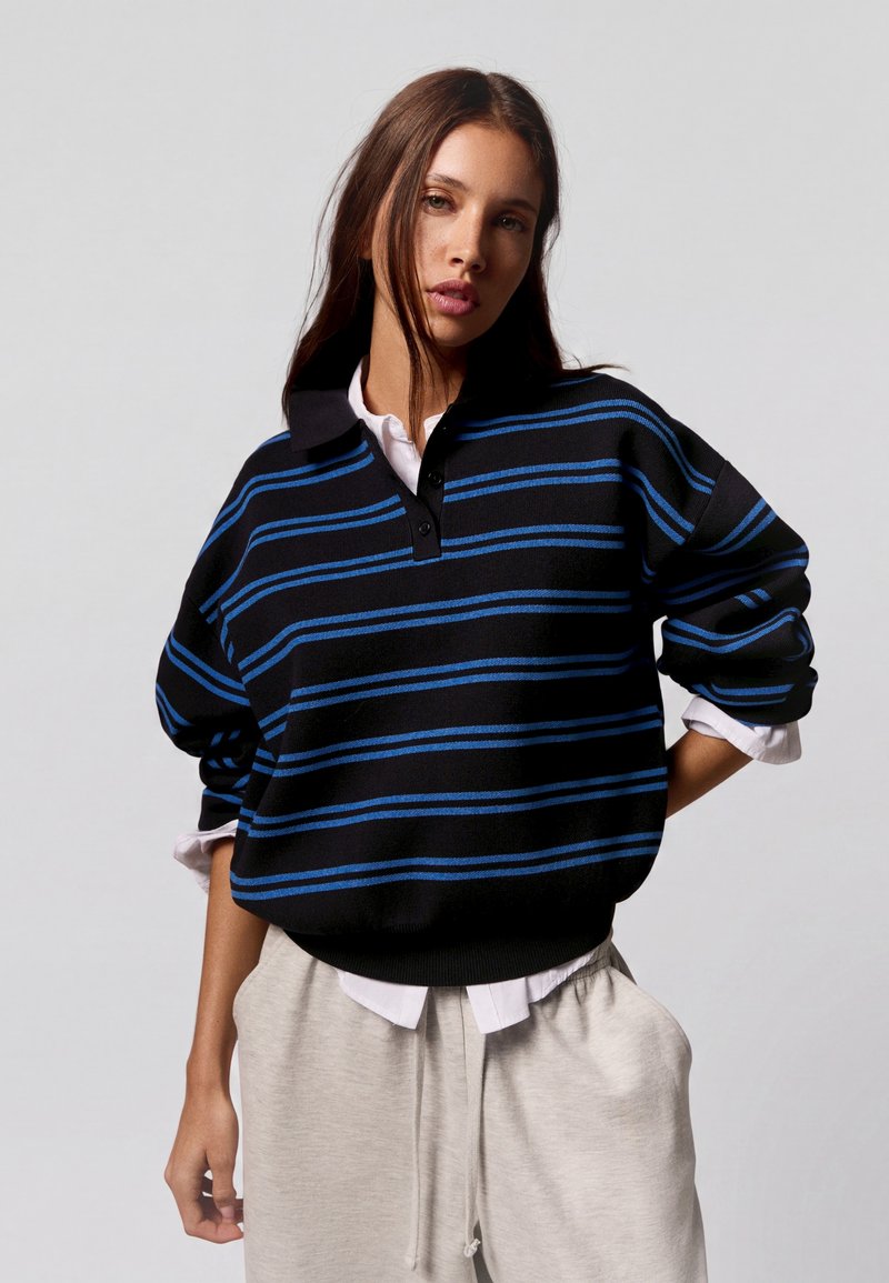 Jeune femme portant un pull rayé noir et bleu sur une chemise blanche et un pantalon beige clair, debout avec une main sur la hanche.