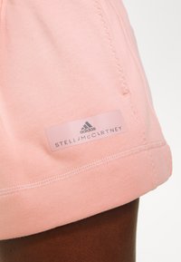 Světle růžové sportovní šortky s jemnou texturou, které mají výraznou značkovou nálepku v elastickém pase. Jsou viditelné detaily šití.