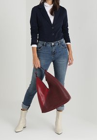 Röd läderhandväska med ett texturerat mönster, triangulär form och släta detaljer; bärs med en marinblå blazer och ljusblå jeans.