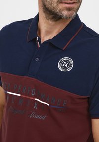 Laivyno mėlynos ir burgundiškos spalvos polo marškiniai su apykakle, trumpomis rankovėmis ir logotipo ženkleliu. Apima teksto akcentus kontrastingomis spalvomis.