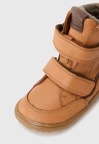 Braune Lederstiefel mit strukturierten Oberflächen, ausgestattet mit zwei Klettverschlüssen und einer abgerundeten Zehenpartie. Innenseite in Braun und Gummisohle.
