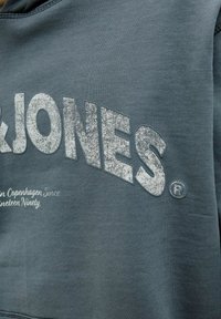 Plavi pamučni dukserica s velikim teksturiranim bijelim natpisom "JONES", uz manje tekstualne i logo detalje u bijeloj boji. Glatka tekstura.