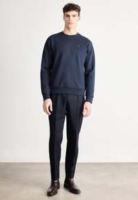 Tmavomodrá mikina s okrúhlym výstrihom a malým logom na hrudi, skombinovaná s tmavými nohavicami na mieru a hnedými koženými čižmami.