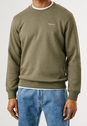 Mies, joka pukeutunut oliivinvihreään pyöreäkaulaiseen collegepaitaan, jossa on pieni valkoinen "Pepe Jeans" -logo, ja vaaleansinisiin farkkuihin, seisoo rennosti käsivarret rentoina.