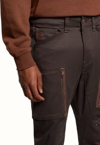 Tummanruskeat työhousut, joissa on useita vetoketjullisia taskuja, teksturoitu kangas ja kontrastivärillä ommeltuja yksityiskohtia. Rennonmalliset, kestävä rakenne.