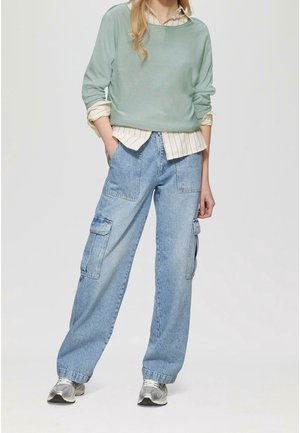 Personne portant un pull vert clair sur une chemise beige rayée, un jean cargo bleu et des baskets argentées, debout les mains dans les poches sur un fond uni.