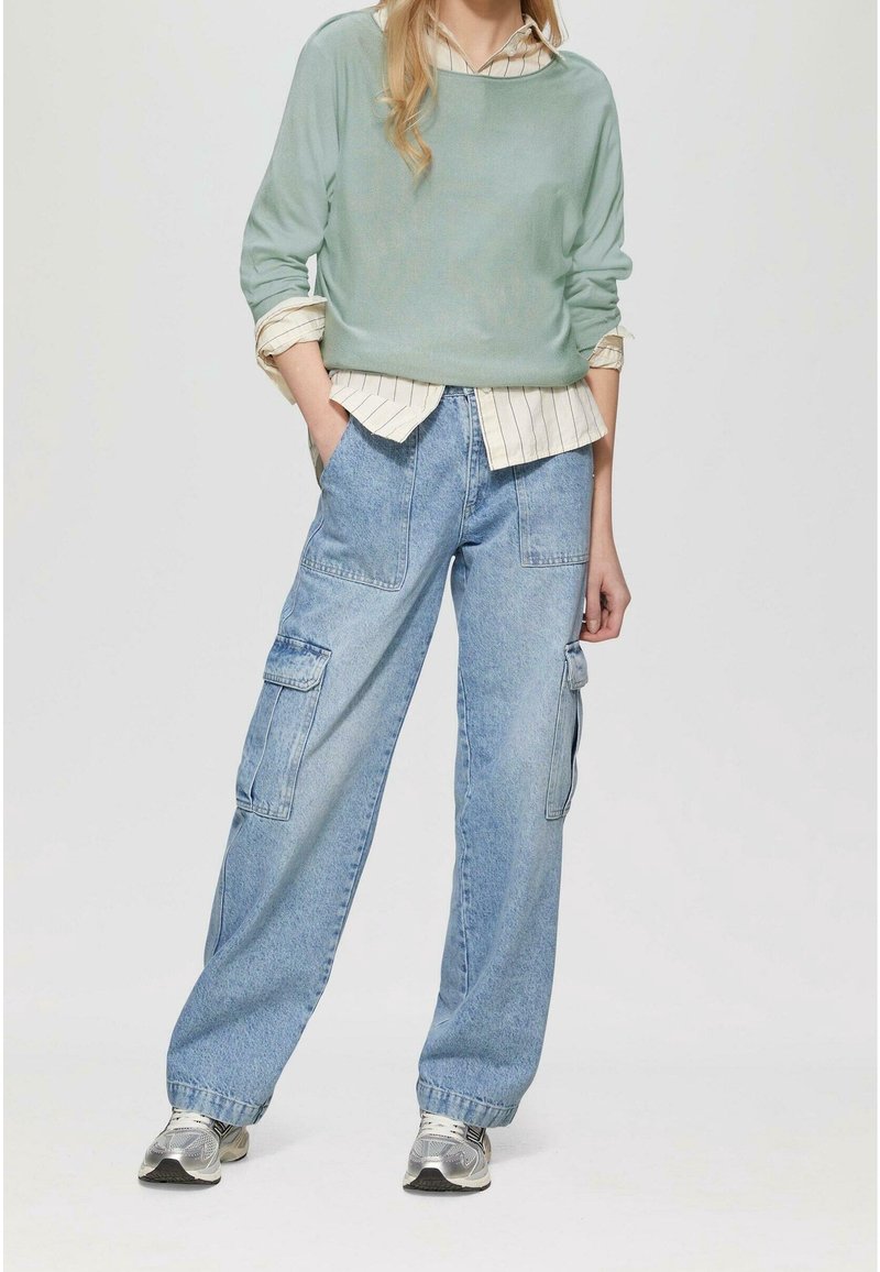 Personne portant un pull vert clair sur une chemise beige rayée, un jean cargo bleu et des baskets argentées, debout les mains dans les poches sur un fond uni.