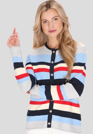 Frau mit langen blonden Haaren, die eine geknöpfte Strickjacke mit horizontalen Streifen in Blau, Rot, Schwarz, Weiß und Grau trägt, steht vor einem einfarbigen Hintergrund.