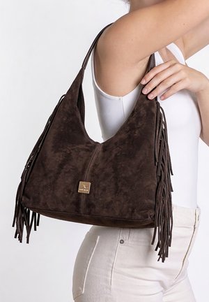 Vrouw met een mouwloos wit topje en lichte broek houdt een grote donkerbruine suède handtas met franjes over haar schouder.