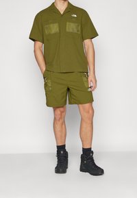 Oliwkowo-zielona koszula z krótkim rękawem z dwoma przednimi kieszeniami z siatki oraz pasujące szorty, oba wykonane z wytrzymałego materiału z akcentami na zamek błyskawiczny. Czarne buty turystyczne.