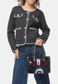 Cardigan in lana nera con bordi bianchi, collo rotondo e bottoni dorati; abbinato a una borsa ricamata nera con motivi colorati e frange.