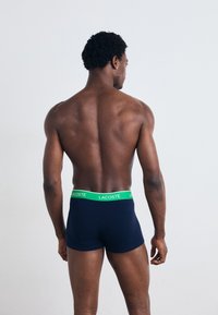 Boxers bleu marine avec un logo vert Lacoste sur la ceinture élastique, vus de l'arrière. Tissu lisse, coupe ajustée.