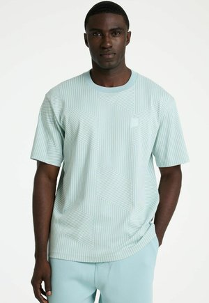 Man met een lichtblauw shirt met korte mouwen en witte geometrische lijnen, gecombineerd met een bijpassende lichtblauwe broek, staand met één hand in zijn zak.