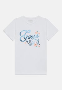 Balta medvilninė marškinėliai su mėlynai blizgančiu "Guess" logotipu ir gėlių akcentais bei tekstu "EST. 1981." Trumpomis rankovėmis ir apvaliu iškirpimu.