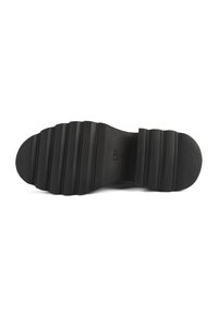 Semelle de chaussure en caoutchouc noir présentant une surface texturée avec des rainures horizontales et un bout arrondi. Logo imprimé près de la zone de la voûte plantaire.