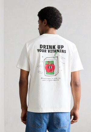 Άνδρας που φοράει λευκό μπλουζάκι με καρτούν πακέτο χυμού μήλου και το κείμενο "Πιες τις βιταμίνες σου" και μπλε τζιν, αντιμετωπίζοντας την κάμερα από την πίσω πλευρά.