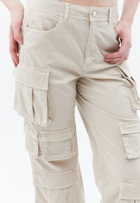 Beige cargo kalhoty s několika bočními kapsami, zipem a knoflíkovým zapínáním, v volném střihu. Vyrobené z lehké tkaniny s minimální strukturou.