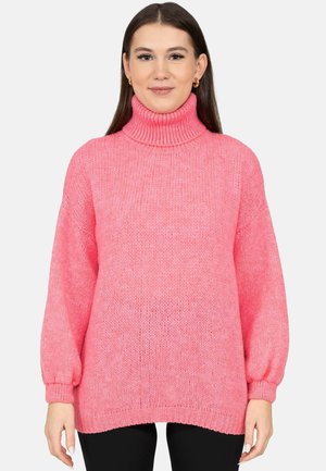 Frau trägt einen hellpink gestrickten Rollkragenpullover mit langen Ärmeln und steht vor einem schlichten hellen Hintergrund.
