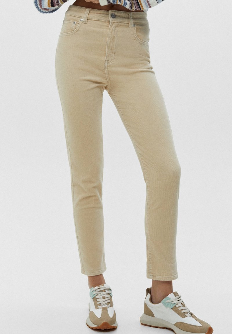 Pantalon en velours côtelé beige slim-fit porté avec des baskets blanches et marron, veste courte visible à la taille, personne debout devant un fond uni.