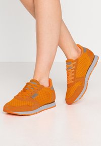 Ljust orange sportskor med mesh- och mockatexturer, med grå accenter. En fot i rörelse mot en enkel vit bakgrund.