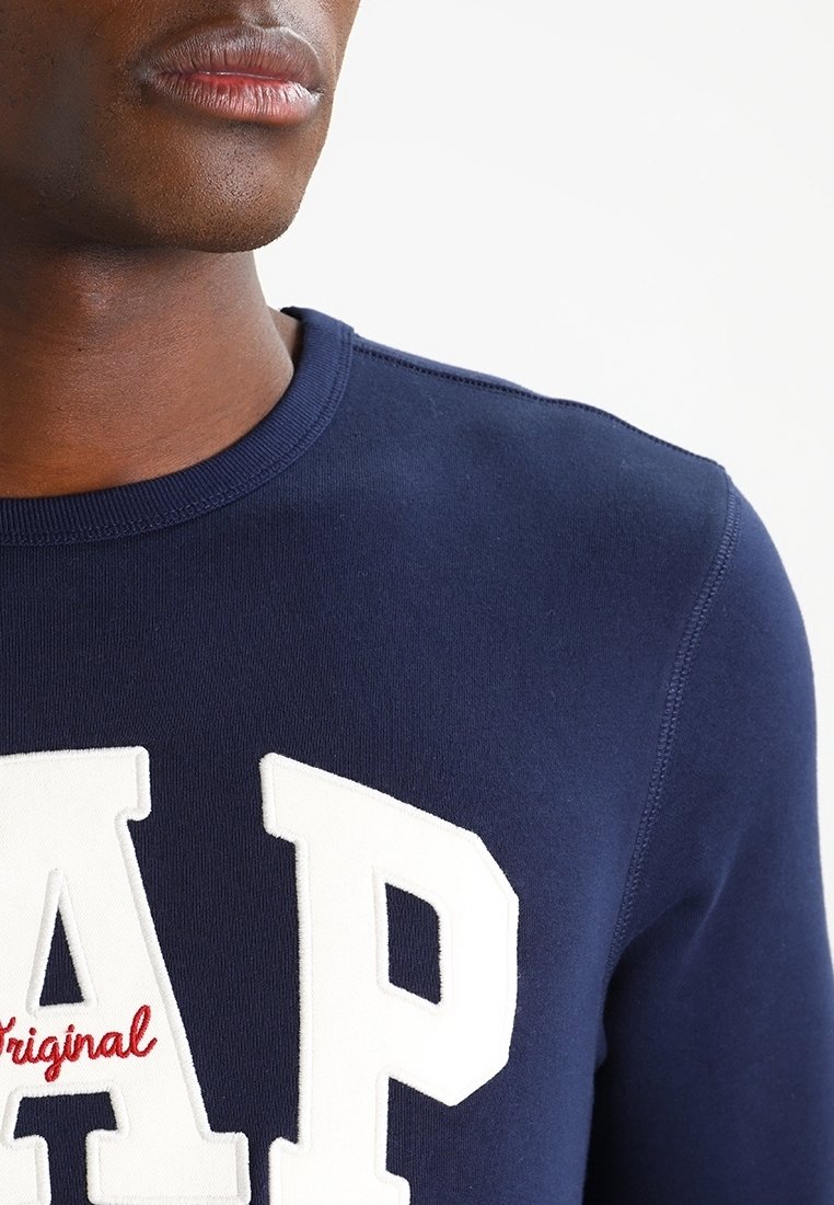 Gros plan d'une personne portant un sweat-shirt bleu marine avec de grandes lettres blanches "AP" et le mot rouge "original" brodé en dessous.