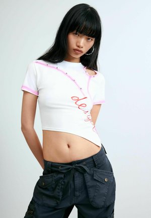 Ung kvinde med langt sort hår iført en hvid kort T-shirt med pink kant og mørke cargobukser mod en ensfarvet baggrund.