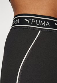 Reggiseno sportivo nero con un'ampia fascia elastica con il marchio Puma, contrastato da una sottile cucitura bianca che corre lungo il lato per un tocco aggiuntivo.