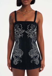 Zwarte denim jurk met dunne bandjes, voorzien van witte geborduurde draken aan de voorkant. Strak aansluitende silhouet en korte zoom.