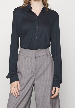 Femme portant une chemise noire à manches longues boutonnée, rentrée dans un pantalon large gris à rayures taille haute.