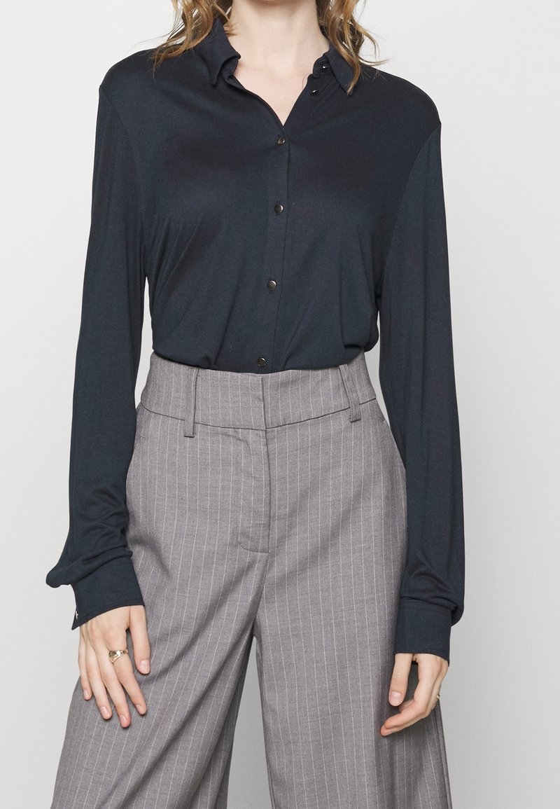 Femme portant une chemise noire à manches longues boutonnée, rentrée dans un pantalon large gris à rayures taille haute.