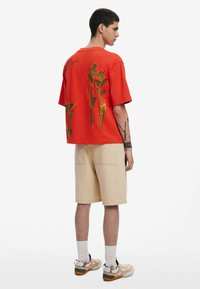 Rød oversized t-shirt med blomstermotiv på ryggen, parret med beige shorts. Outfittet har en afslappet pasform og et enkelt design.