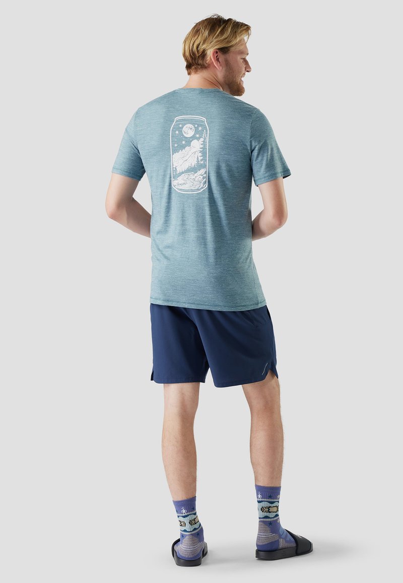 Uomo che indossa una T-shirt grafica verde acqua con disegno di montagna e luna, pantaloncini blu scuro, calzini viola fantasia e sandali neri a fascia, in piedi di spalle.