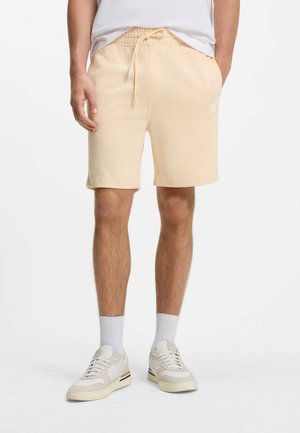 Mann trägt hellbeige Shorts mit Kordelzug, weiße Sneakersocken und weiße Low-Top-Sneaker, steht vor einem schlichten weißen Hintergrund.